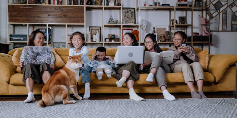 A Family Sitting On Sofa, The Kids Playing With A Dog, And Adults Integrating Asset Protection Trusts In Their Estate Plan With Their Laptop.